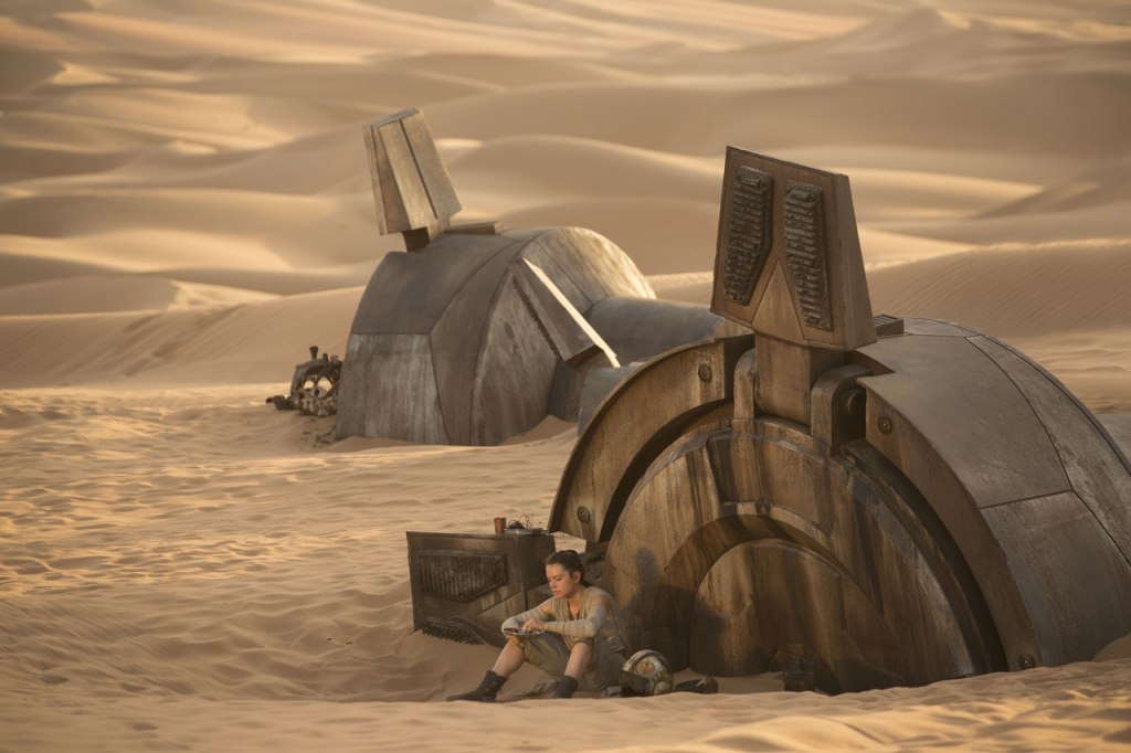 Screenshot from The Force Awakens: Rey sits outside in the desert at sunset next to the toppled ruins of a metal armature.